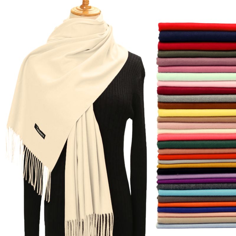 Beige scarf with fringe draped over a black sweater, next to a stack of folded scarves in various colors.