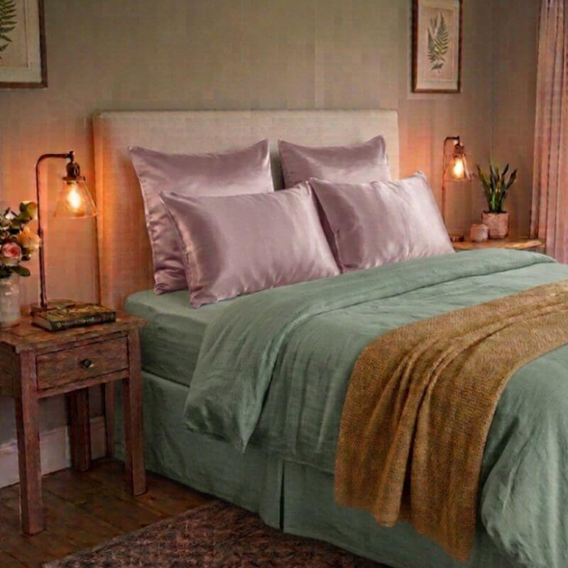 A styled bedroom scene showing a bed made up with sage green bedding, matching silk pillows, and a tan throw blanket. The bed is set in a warm, dimly lit room with wooden nightstands, bedside lamps, and framed wall art.