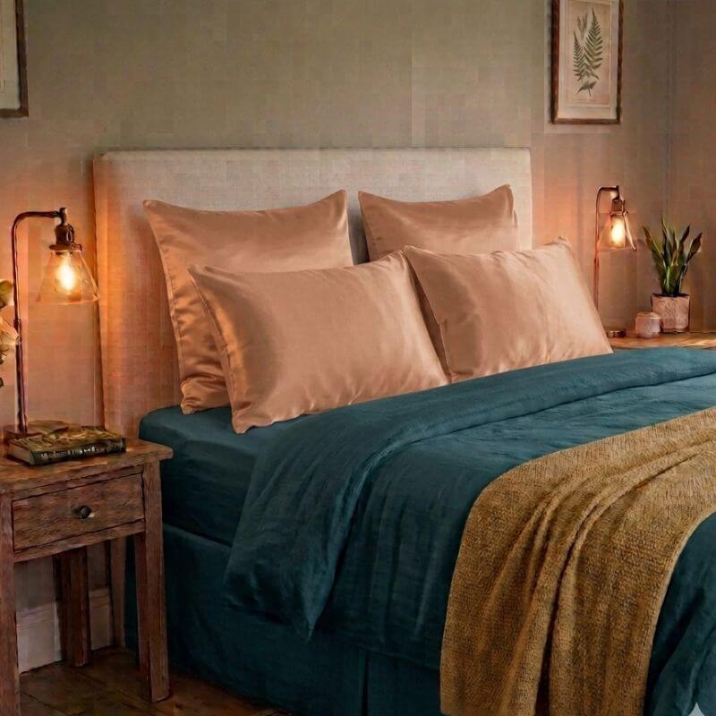 A neatly made bed in a warmly lit bedroom, styled with a teal duvet cover, gold silk pillowcases, a beige upholstered headboard, and a tan throw blanket. A wooden bedside table with a lit table lamp sits on the left.